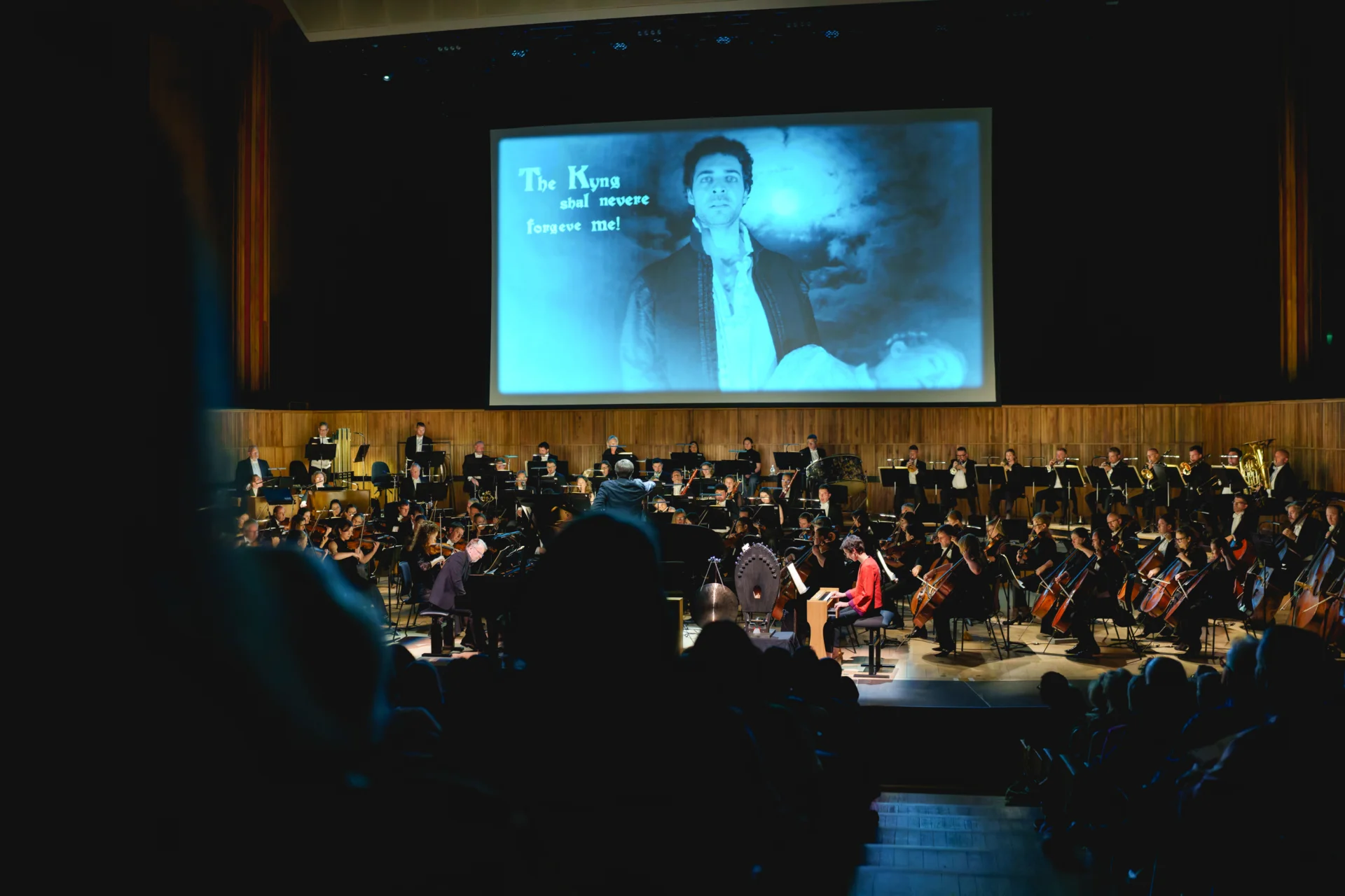 The RPO performing on stage viewed from behind audience members in the stalls. On the screen behind the Orchestra it shows a man with a cyanotype coloured filter