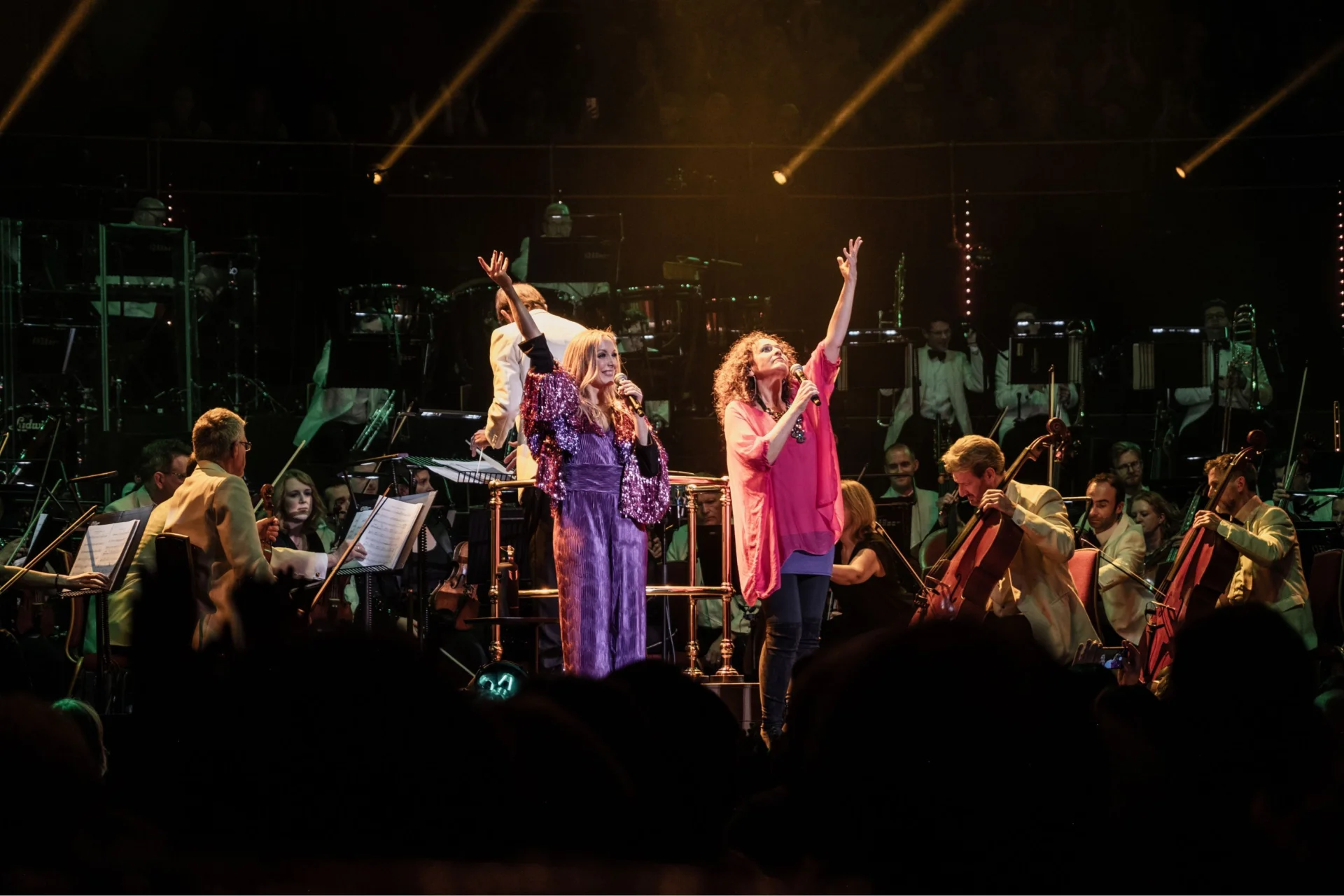 Two ABBAphonic vocalists stand on stage at the Royal Albert Hall in front of the Orchestra, arms outstretched