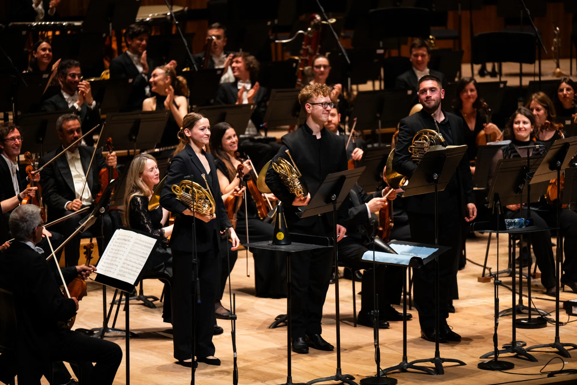 Katy Woolley, Alexander Edmundson and Ben Hulme standing at the front of the stage holding their horns with the orchestra applauding behind them