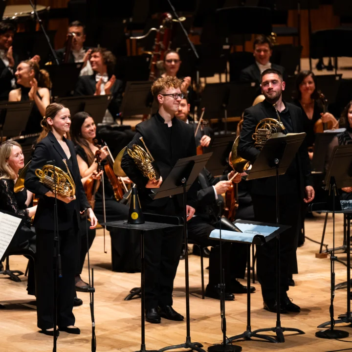 Katy Woolley, Alexander Edmundson and Ben Hulme standing at the front of the stage holding their horns with the orchestra applauding behind them