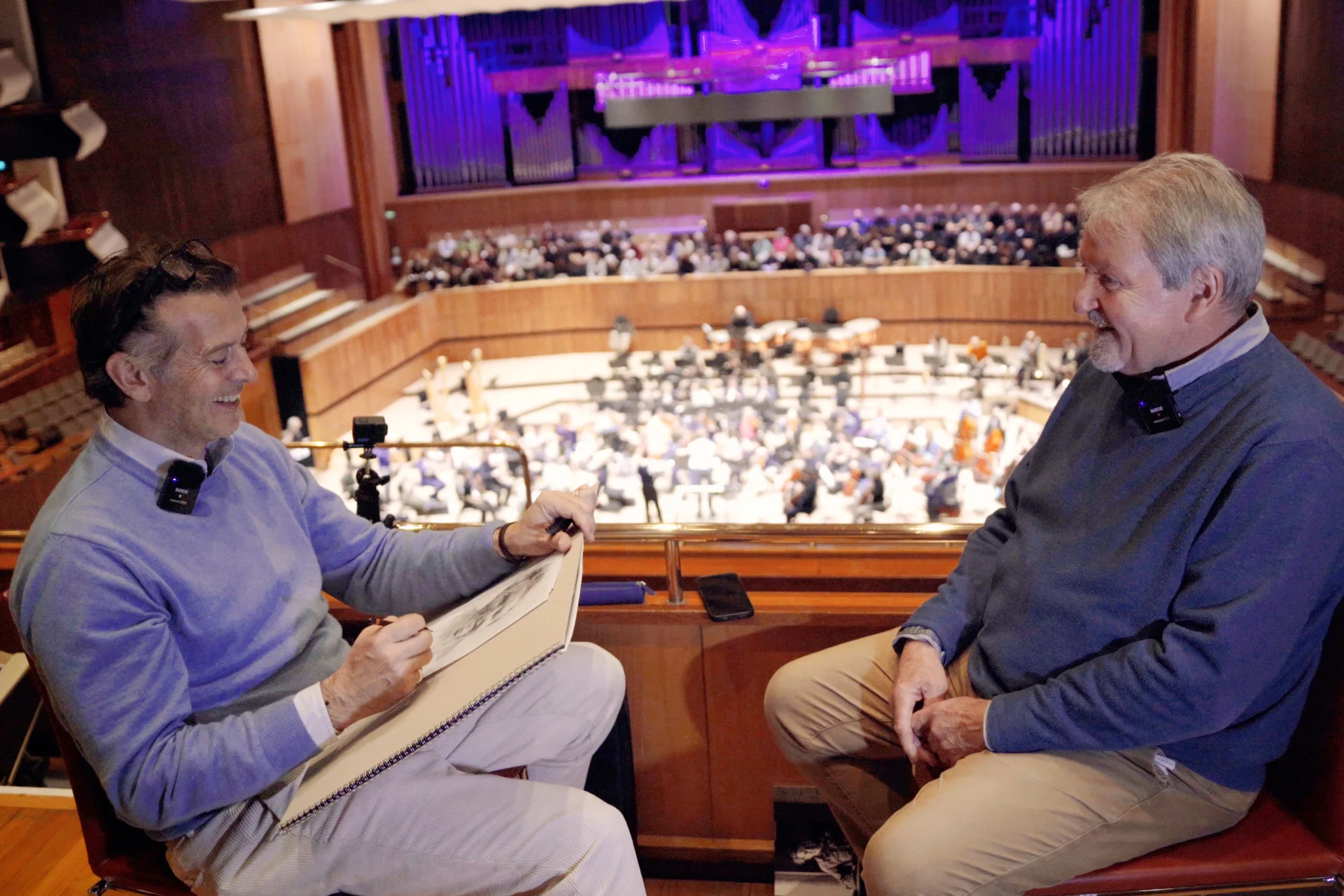 Artist Jeremy Houghton sketching violinist Andrew Klee in the upper stalls of the Royal Festival Hall with the Orchestra and a choir getting ready to rehearse on stage in the background