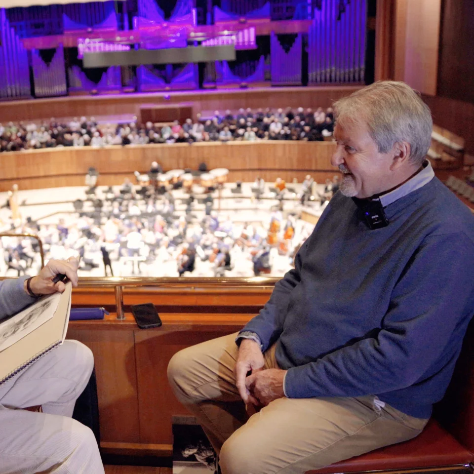 Artist Jeremy Houghton sketching violinist Andrew Klee in the upper stalls of the Royal Festival Hall with the Orchestra and a choir getting ready to rehearse on stage in the background