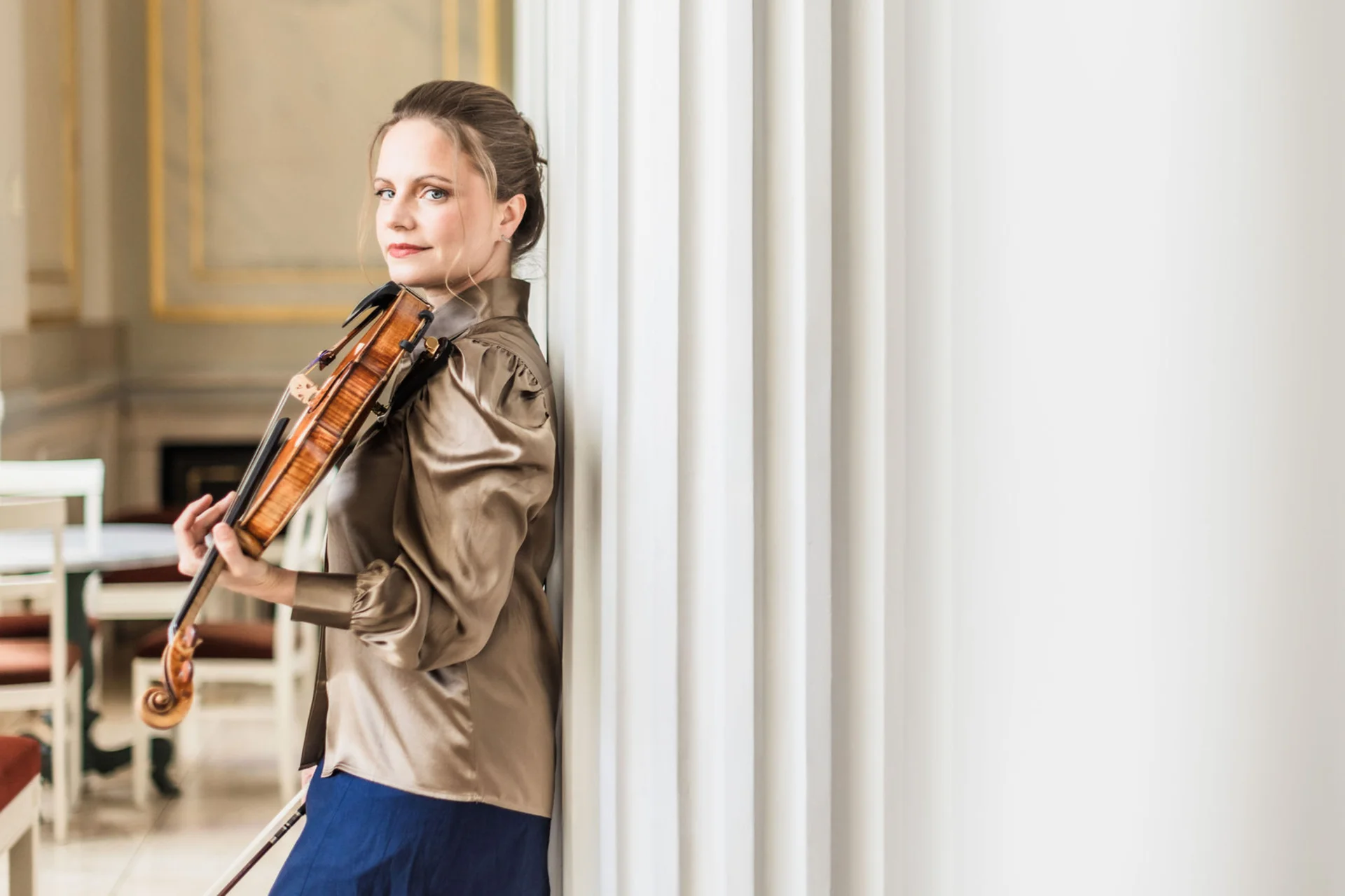 Julia Fischer leaning against a pillar with her violin and looking at the camera