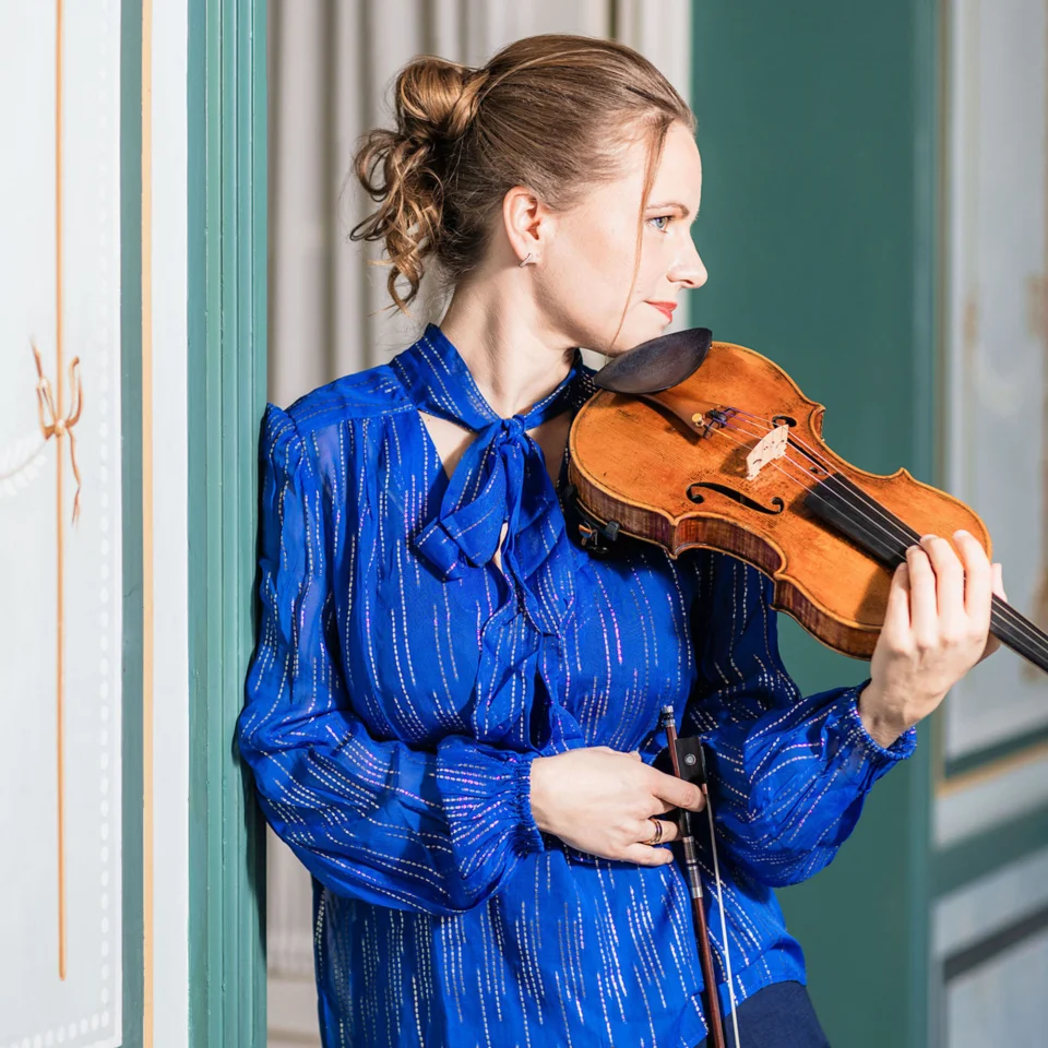 Julia Fischer in a blue top and holding her violin looking away from the camera