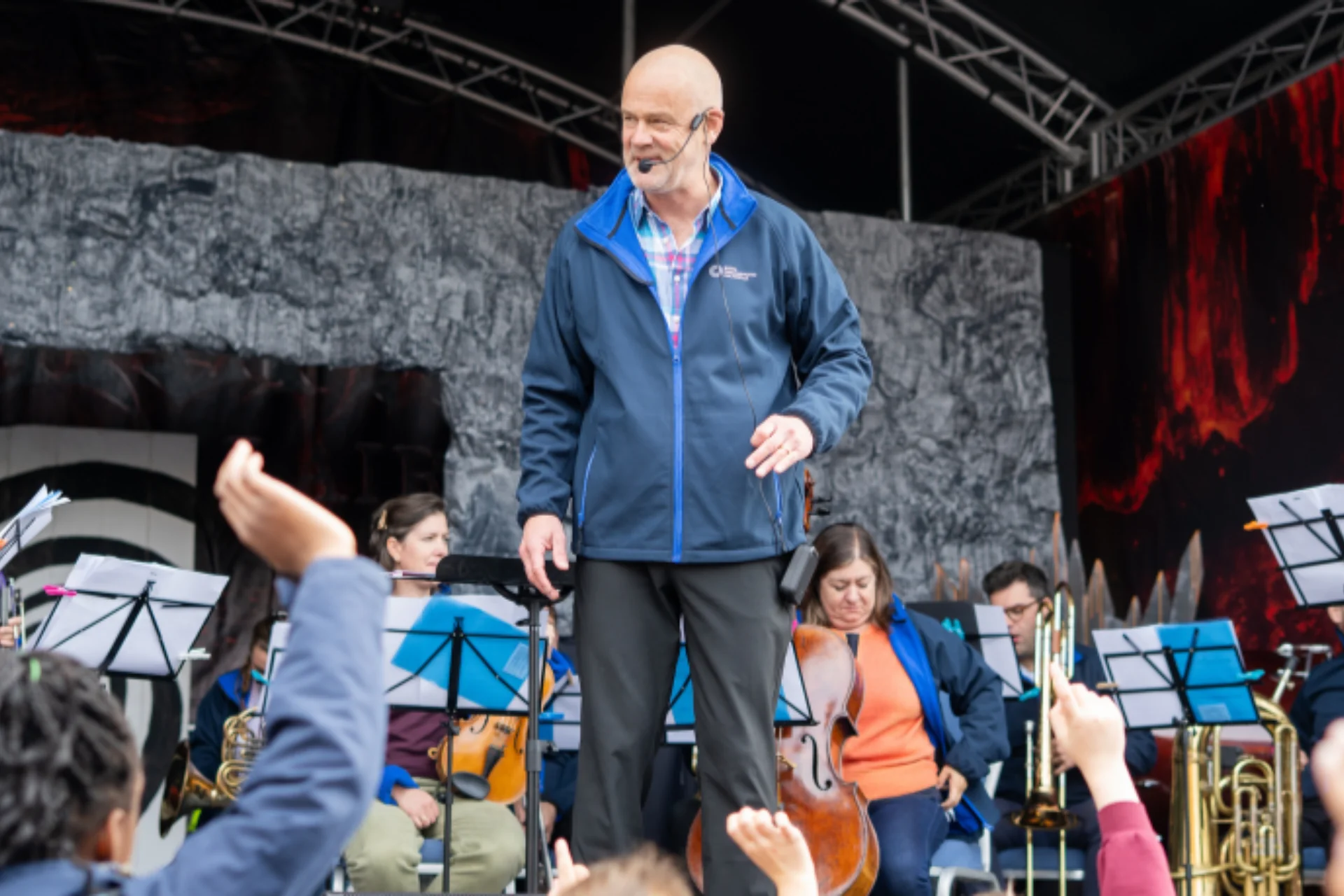 Conductor and Orchestra on stage, young people with hands raised to participate