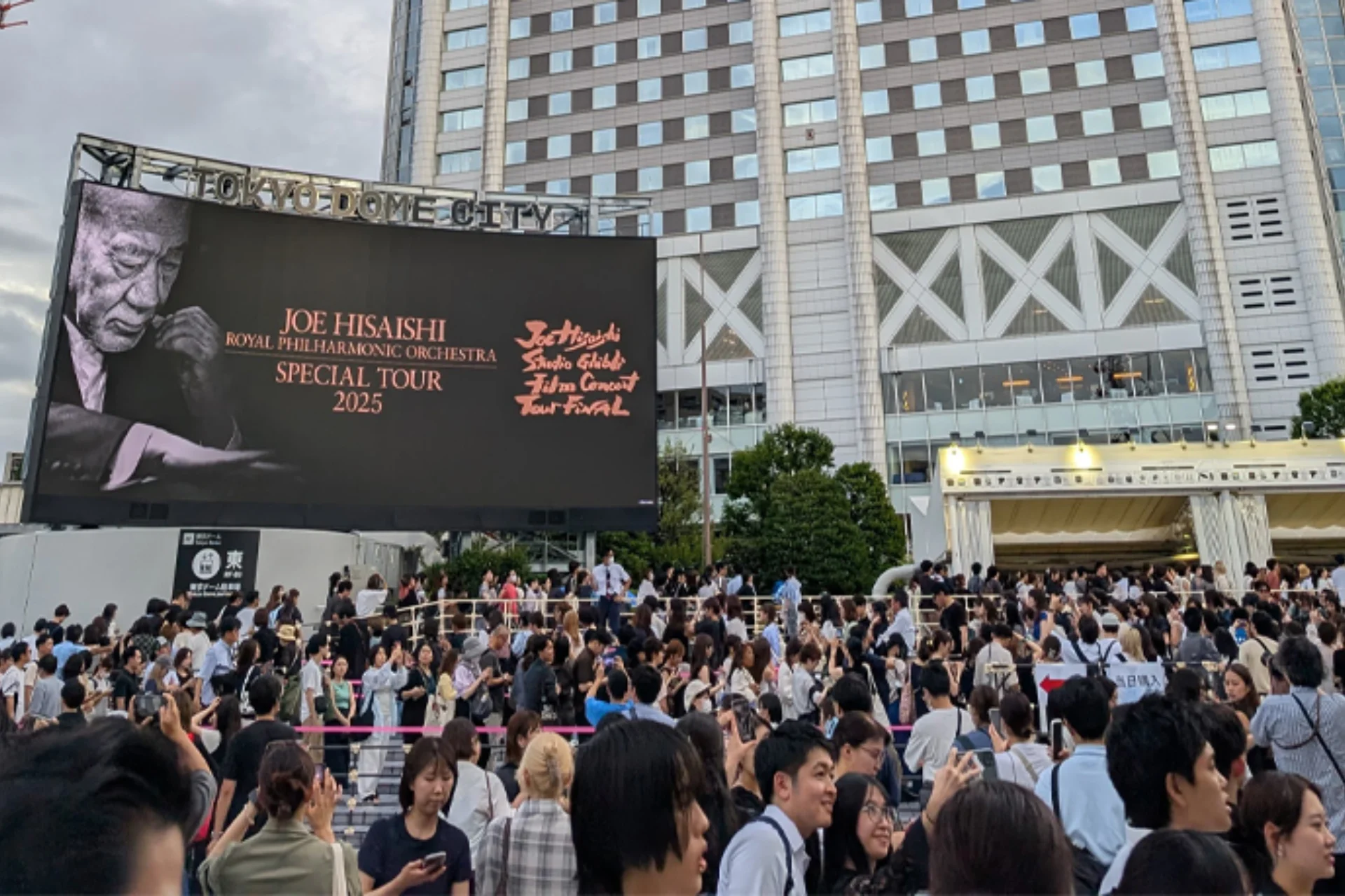 Queues of people outside Tokyo Dome with a billboard above them advertising the Joe Hisaishi and RPO Studio Ghibli music film tour