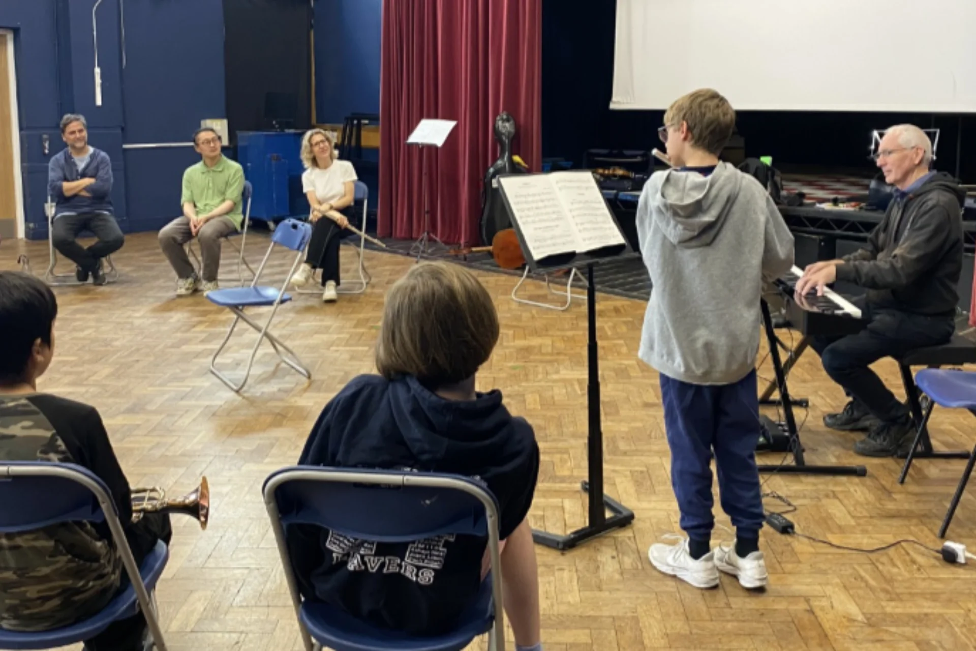 A boy performing in front of a music stand with a keyboard accompanist, as others watch in a school hall.