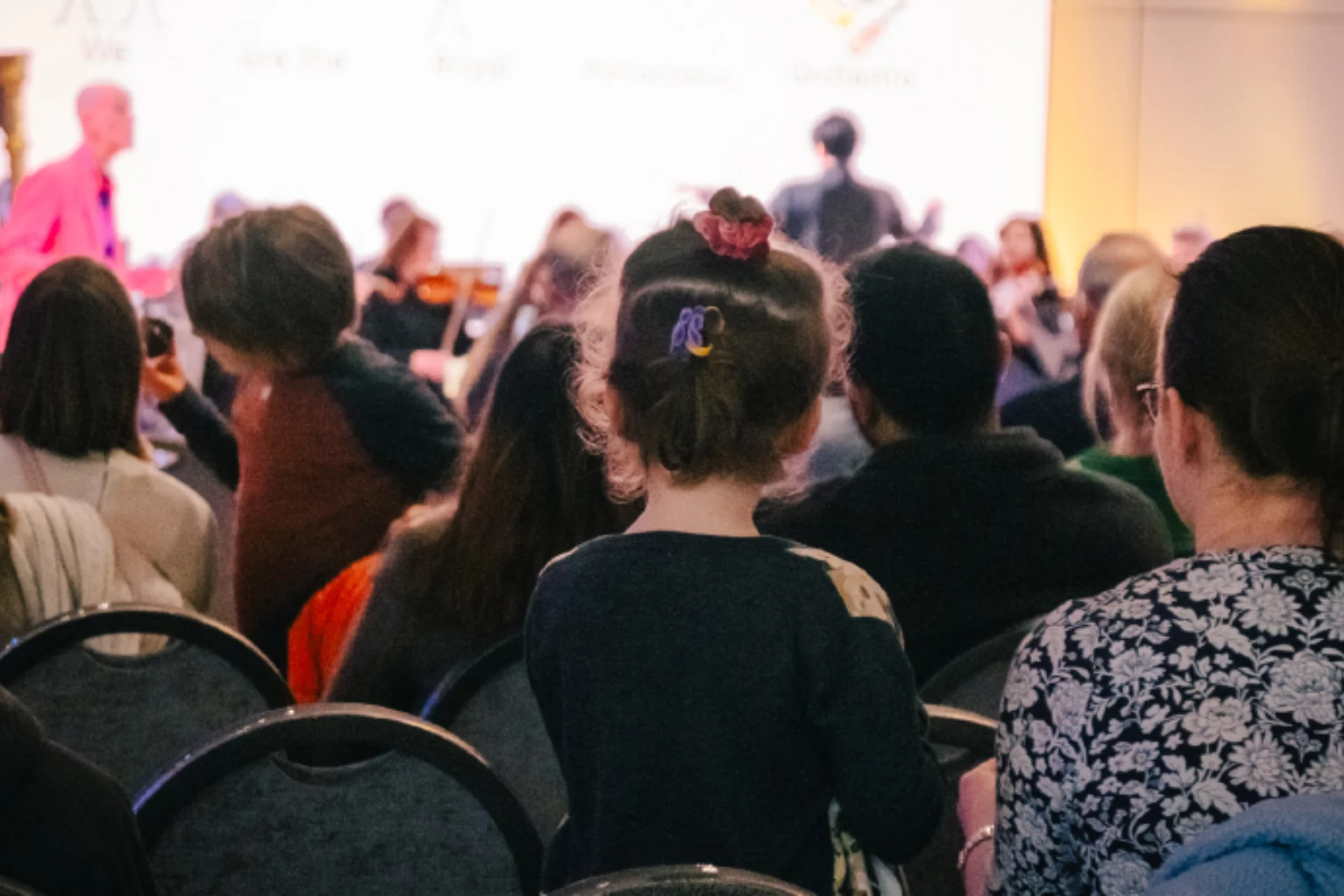 An audience of children and adults watching the RPO perform in Brent Civic Centre, with a presenter in a pink suit standing in front of the Orchestra