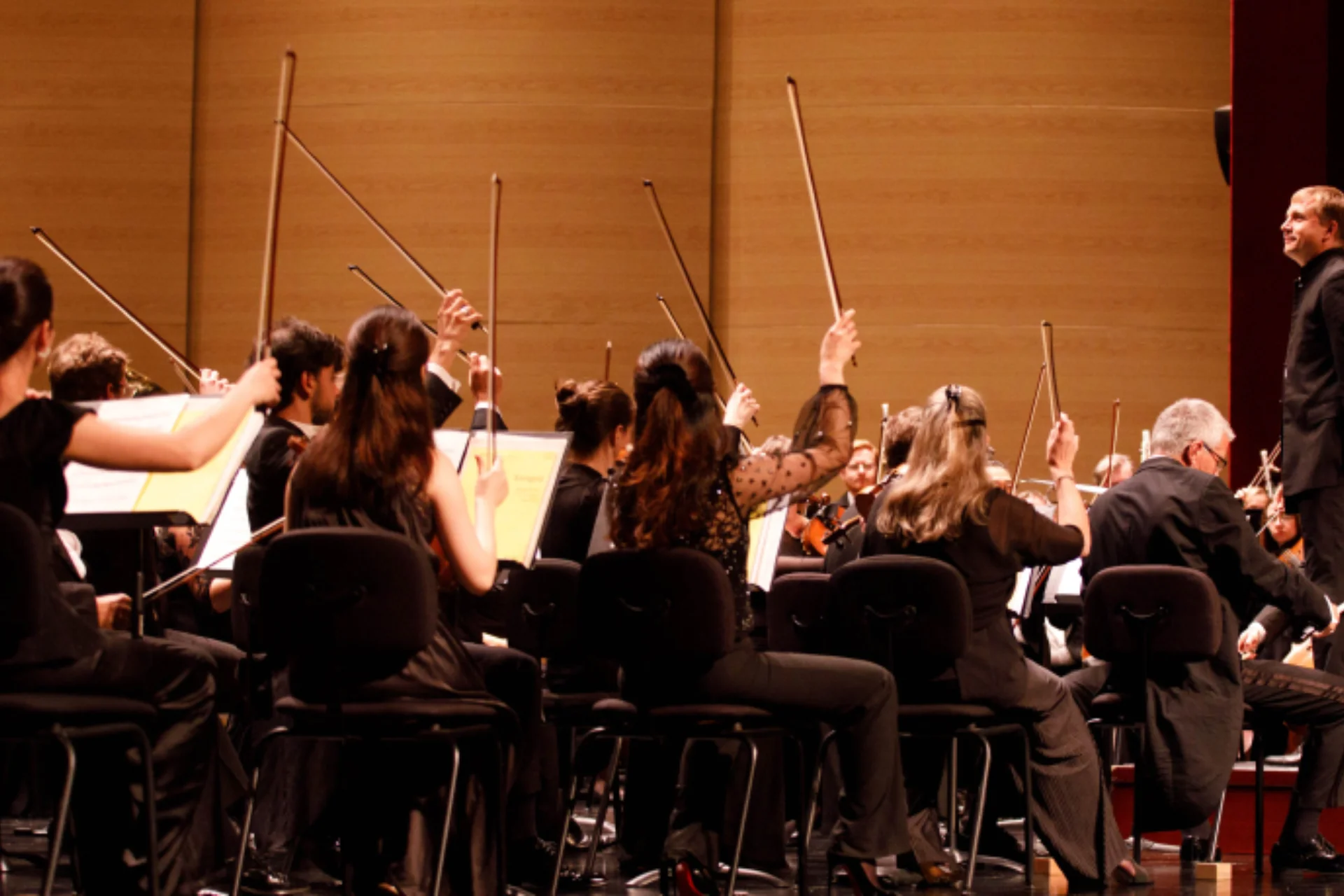 The first violins raising their bows in the air after playing a final note, and conductor Vasily Petrenko standing on the podium and smiling