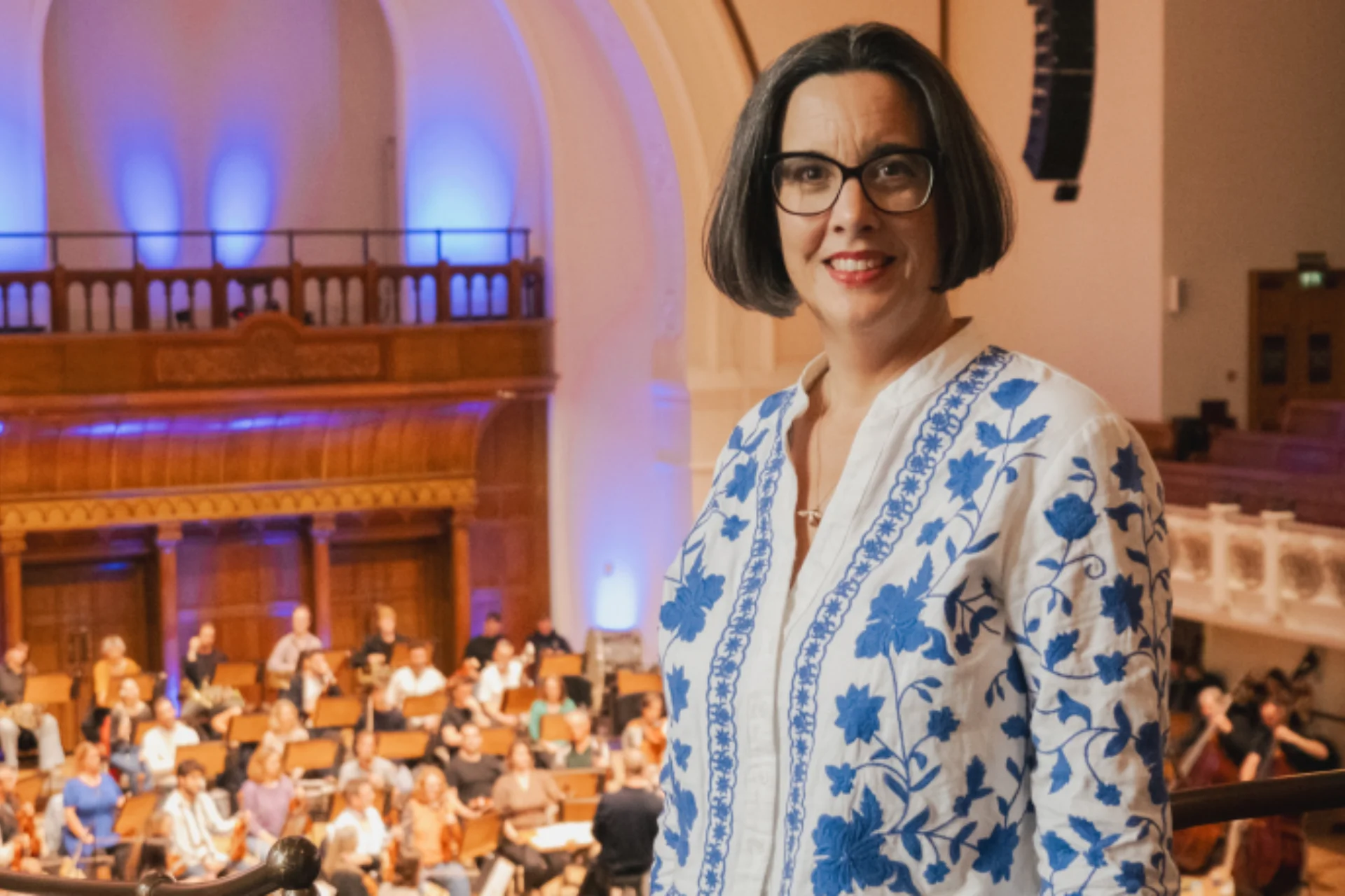 Sarah Bardwell in  white cardigan will blue flowers standing and smiling on the upper Gallery at Cadogan Hall, with the Orchestra rehearsing behind her