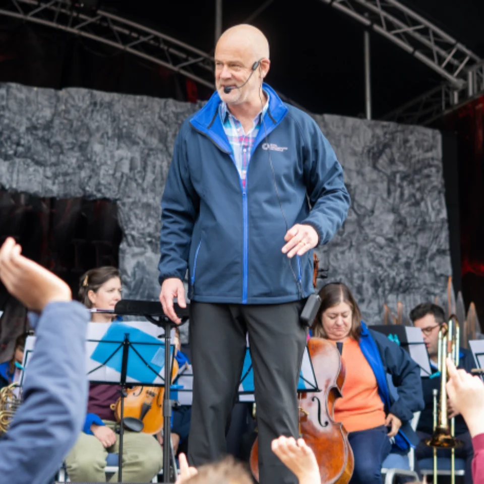 Conductor and Orchestra on stage, young people with hands raised to participate
