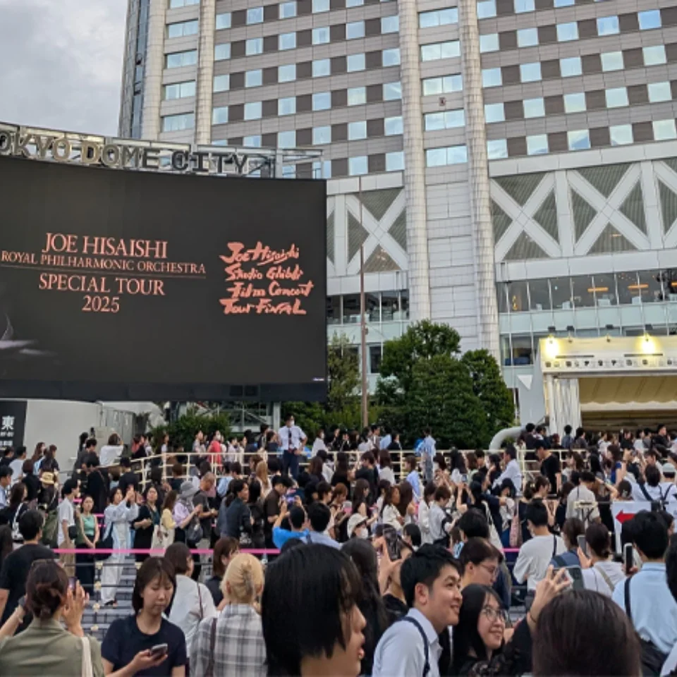 Queues of people outside Tokyo Dome with a billboard above them advertising the Joe Hisaishi and RPO Studio Ghibli music film tour