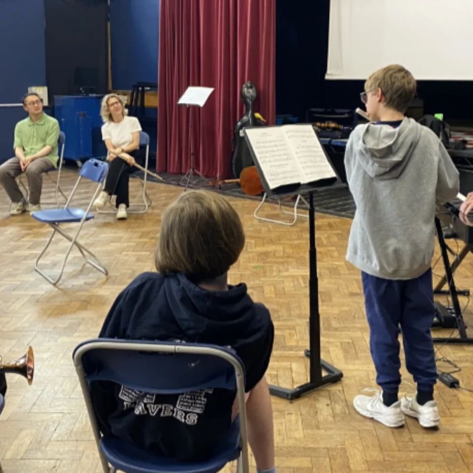 A boy performing in front of a music stand with a keyboard accompanist, as others watch in a school hall.