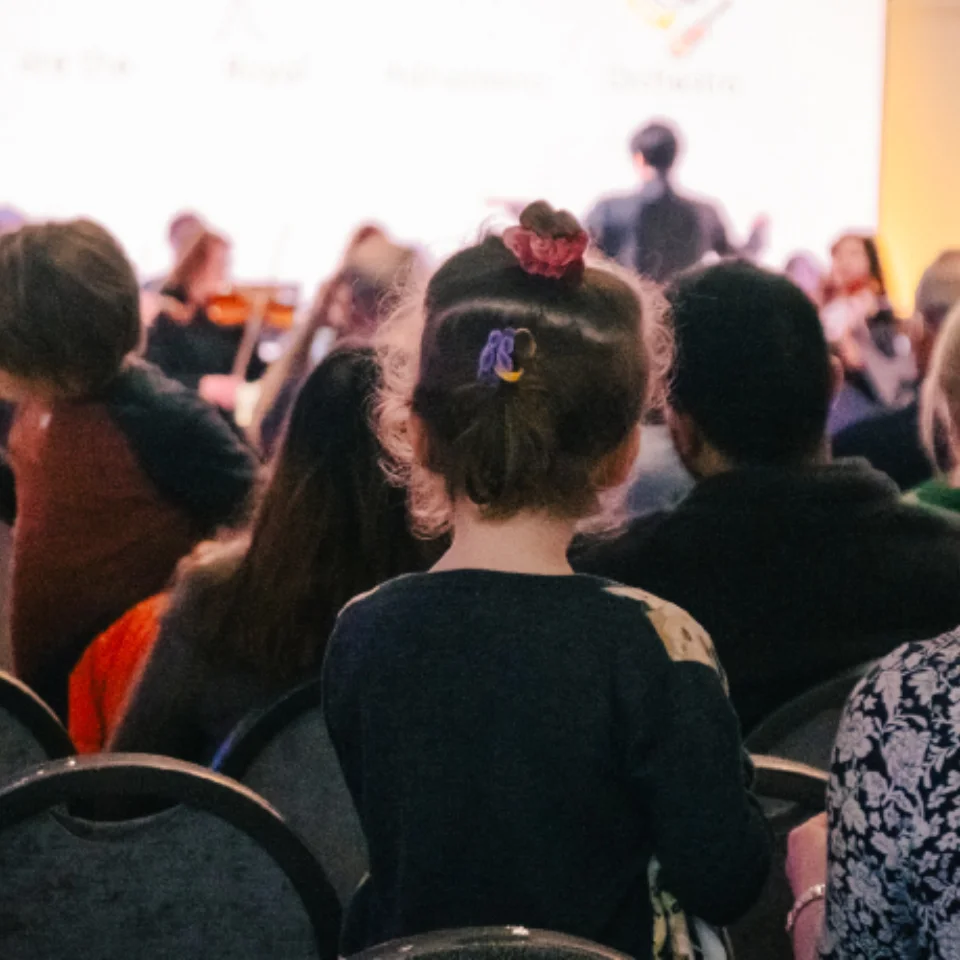 An audience of children and adults watching the RPO perform in Brent Civic Centre, with a presenter in a pink suit standing in front of the Orchestra