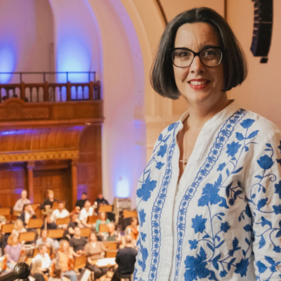 Sarah Bardwell in  white cardigan will blue flowers standing and smiling on the upper Gallery at Cadogan Hall, with the Orchestra rehearsing behind her