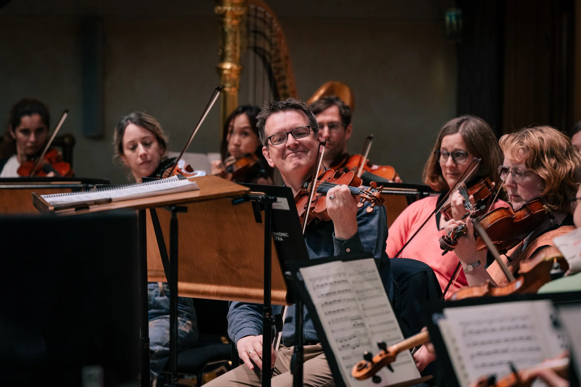 A close up shot of the strings section of the Royal Philharmonic Orchestra rehearsing at Cadogan Hall. One violinist in the center smiles as he plays his violin.