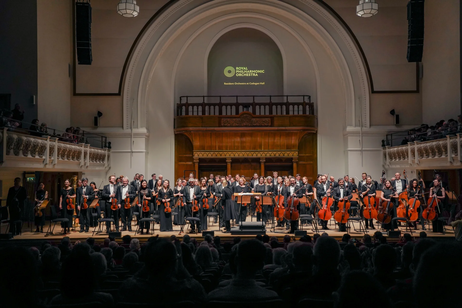 The Royal Philharmonic Orchestra stands for applause onstage at Cadogan Hall. Above them is a rounded archway and a projection of the Royal Philharmonic Orchestra logo