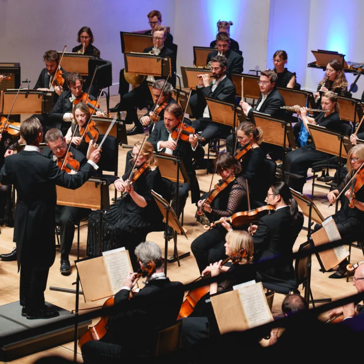 A conductor stands with his arms raised in front of the string section of the Royal Philharmonic Orchestra, performing at Cadogan Hall
