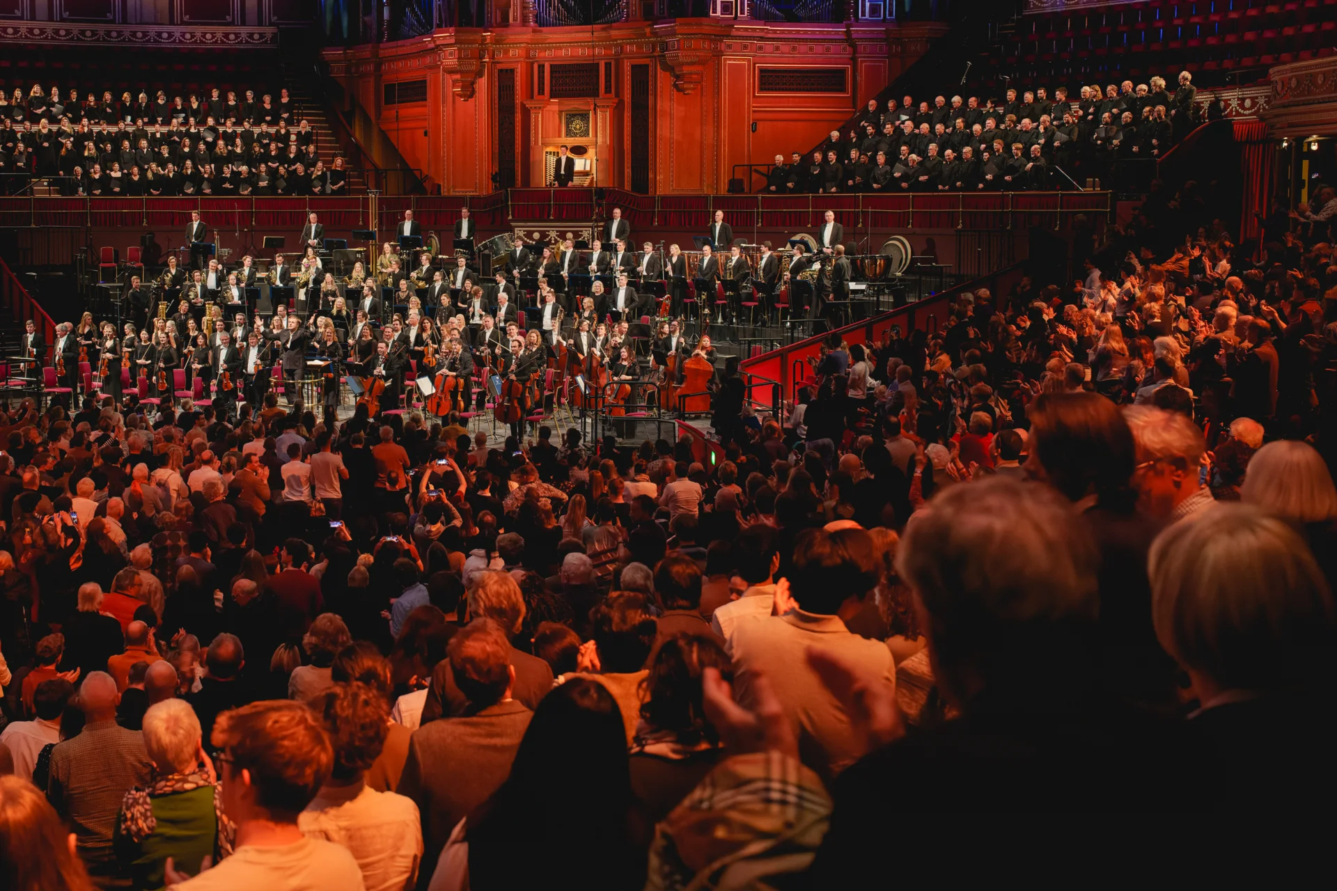 Audience members standing to applause as the Orchestra and a choir stands on stage and in the choir stalls at the Royal Albert Hall