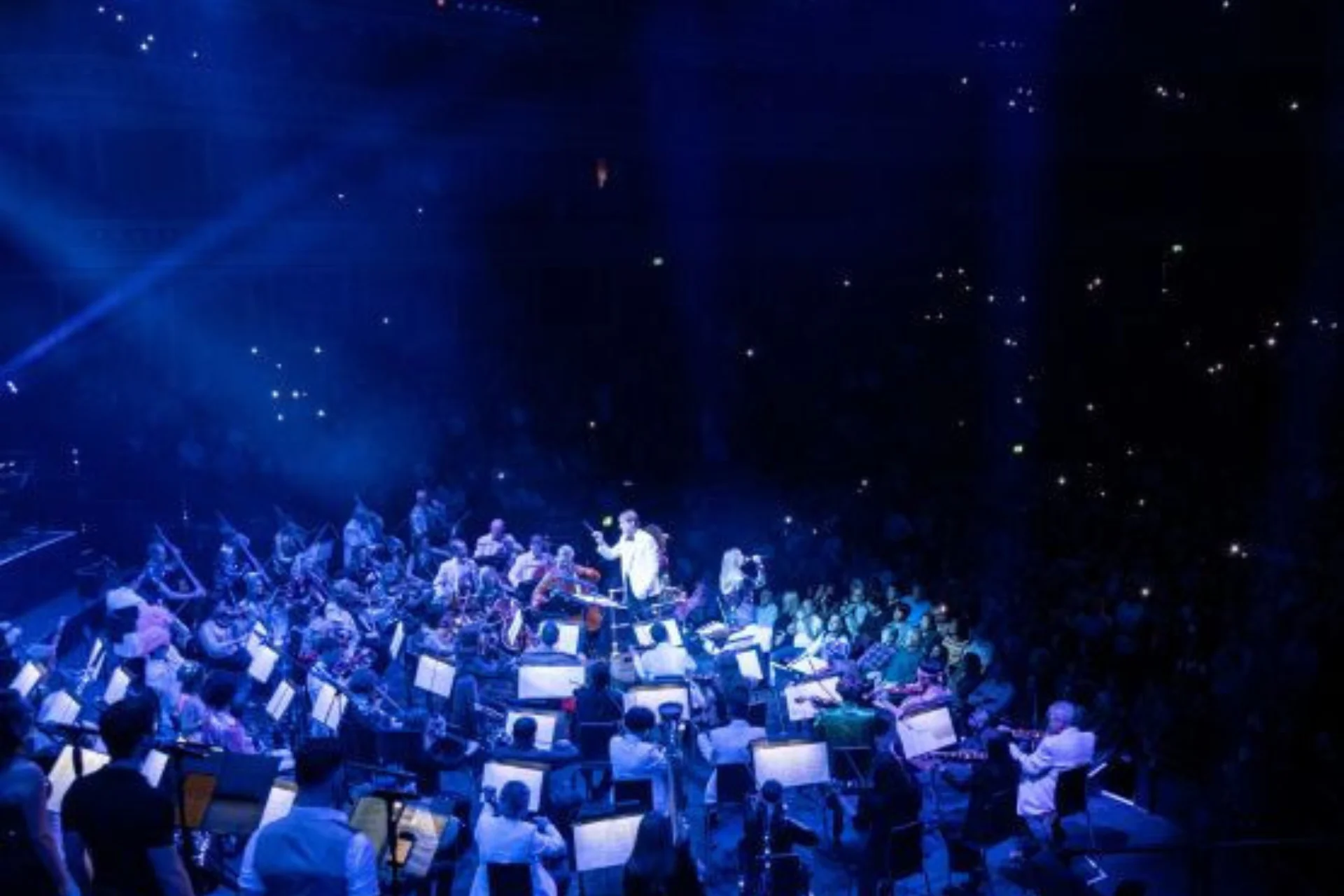 The Royal Philharmonic Orchestra on stage at the Royal Albert Hall. They are engulfed in blue stage lights, and the audience shimmers in the background.