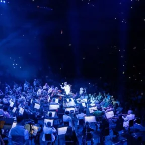 The Royal Philharmonic Orchestra on stage at the Royal Albert Hall. They are engulfed in blue stage lights, and the audience shimmers in the background.