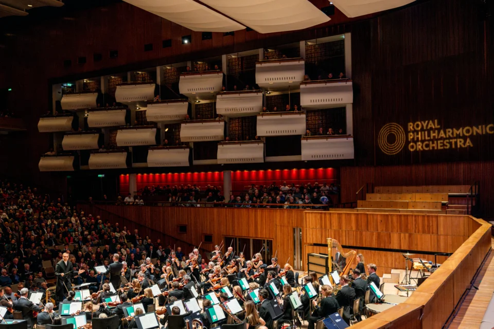 The Royal Philharmonic Orchestra onstage at the Royal Festival Hall, with the audience stretching in front of them. The Royal Philharmonic Orchestra logo is projected onto the wall nearby.