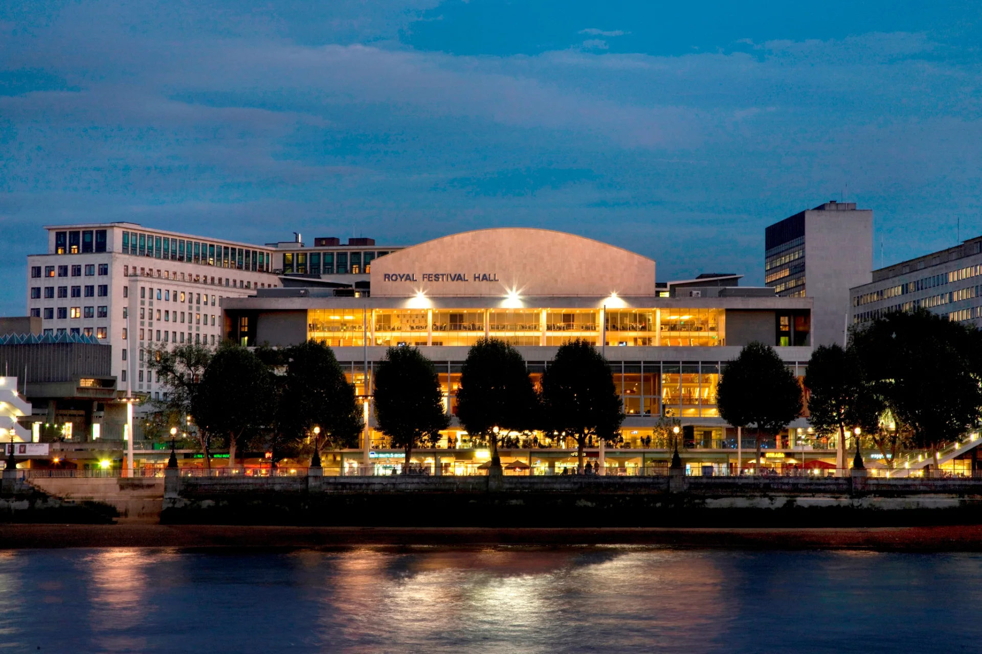 View of the Royal Festival Hall from across the Thames River at dusk. The lights of the venue reflect off the water.