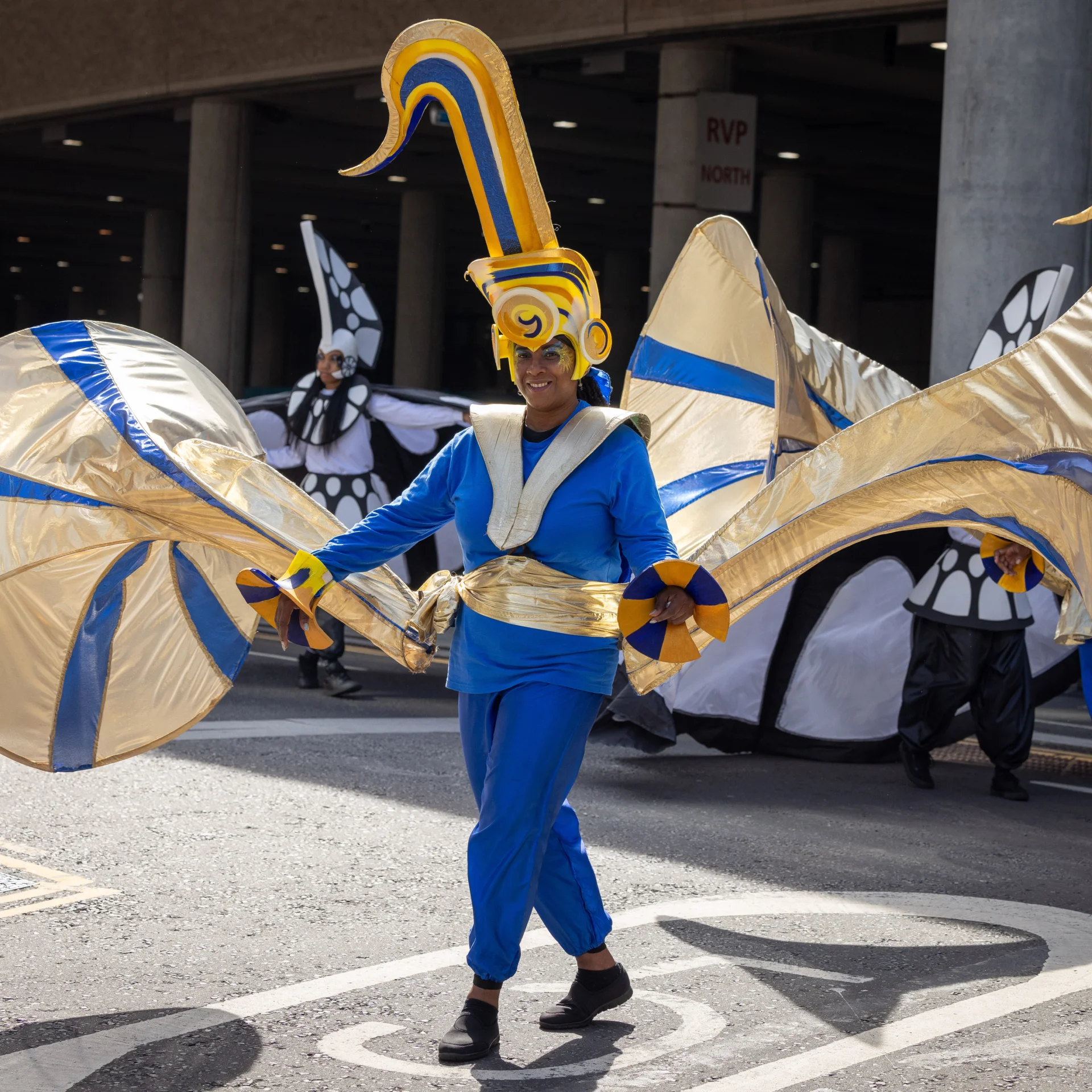 A person is in a blue and white carnival costume with trumpet bells as big as a person coming out of each of their arms and a saxophone on their head.