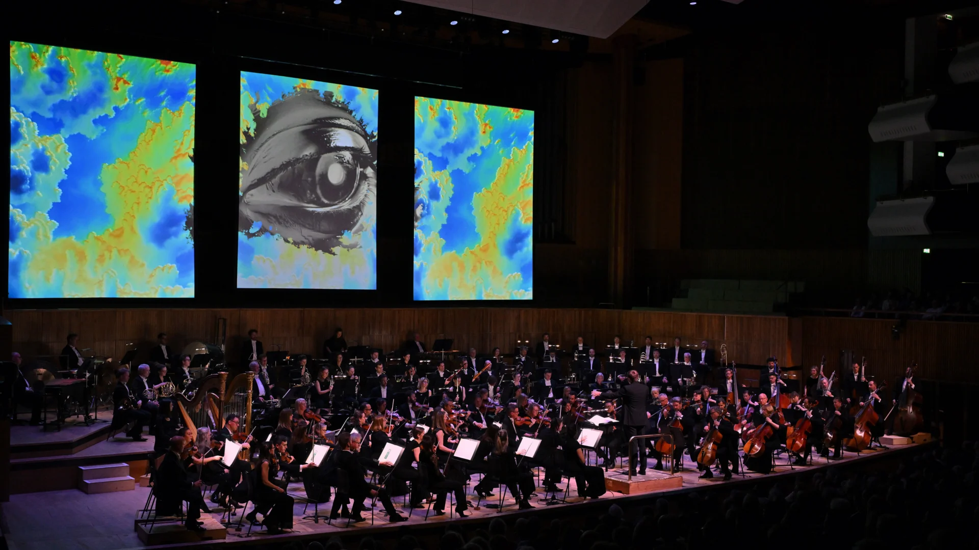 The RPO conducted by Vasily Petrenko on stage at the Royal Festival Hall, with three screens behind the stage showing a blue and yellow cloudy artwork with a huge silver eye in the centre