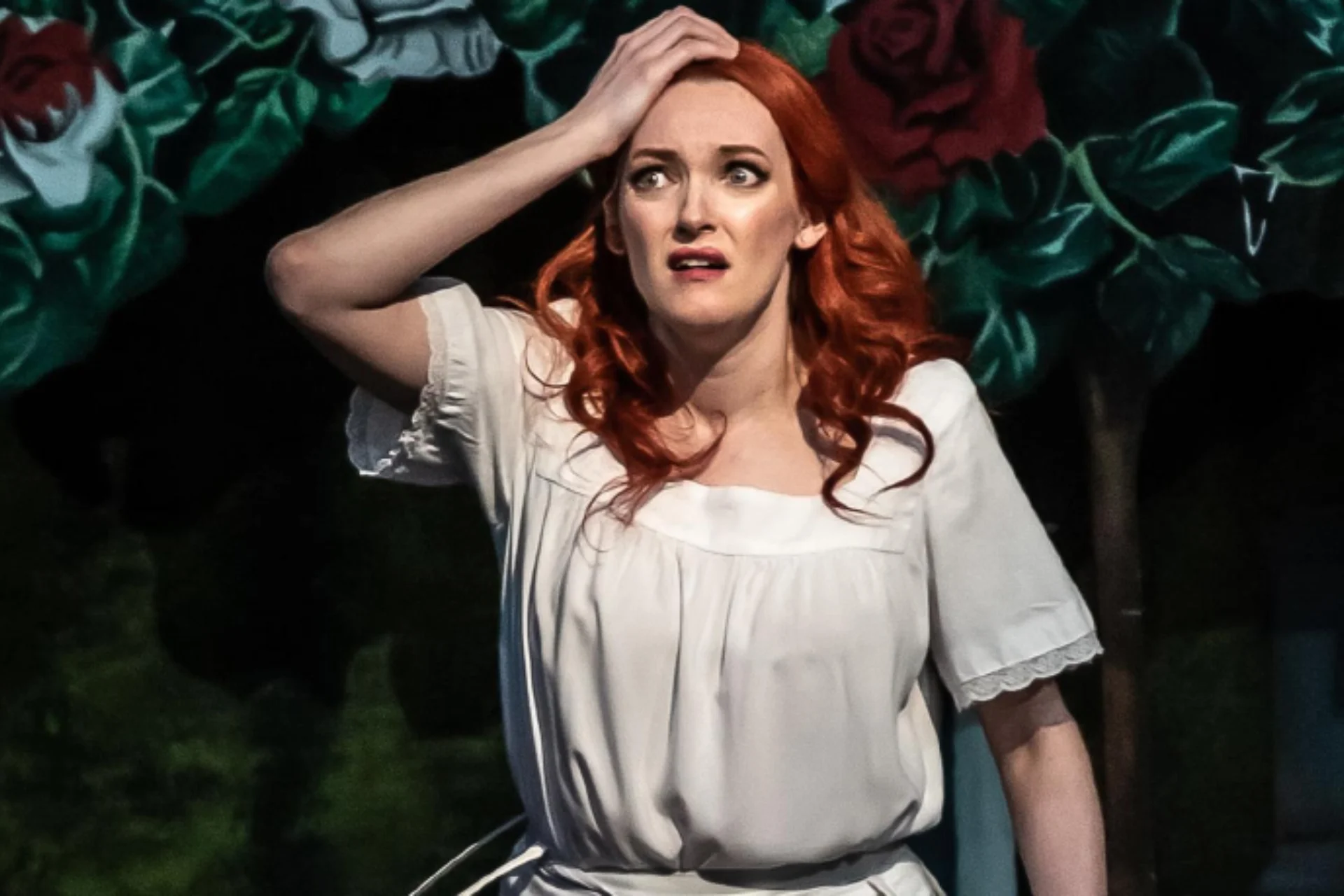 Soprano Jennifer France in a frilled white shirt holding her hand to her head in front of an opera scene of roses and greenery