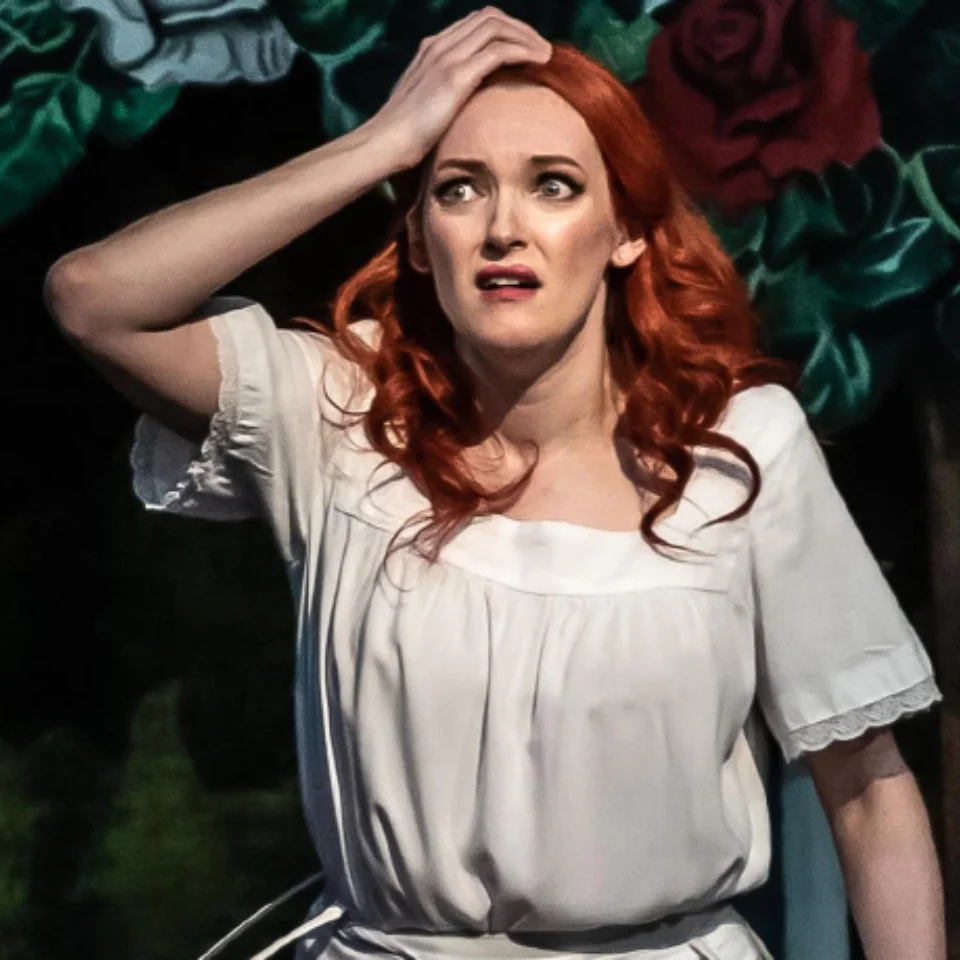 Soprano Jennifer France in a frilled white shirt holding her hand to her head in front of an opera scene of roses and greenery