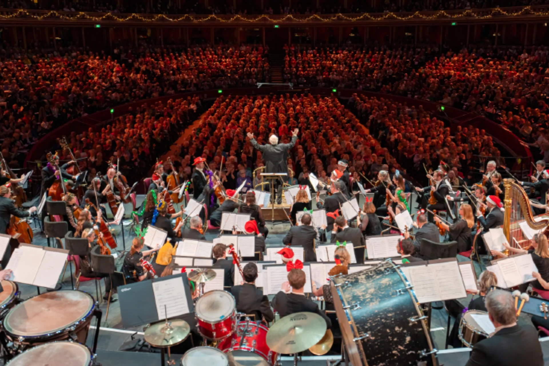 John Rutter conducting the audience form the podium at the Royal Albert Hall, with the Orchestra wearing Christmas accessories