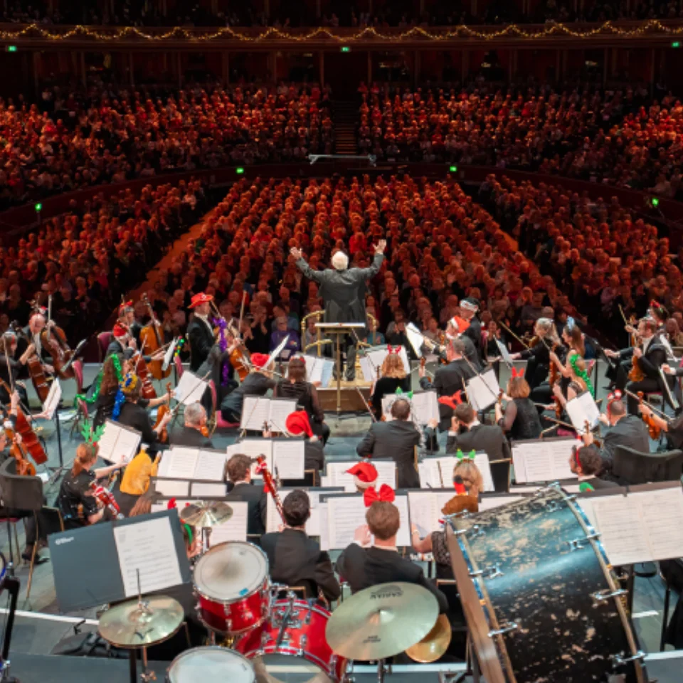 John Rutter conducting the audience form the podium at the Royal Albert Hall, with the Orchestra wearing Christmas accessories